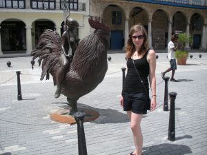Holly Roose posing next to a statue of a rooster with a nude woman riding it and holding a fork nearly the same size of her body